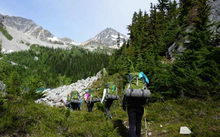 a group of people carrying backpacks hike through a green clearing. There are mountains in the background. 